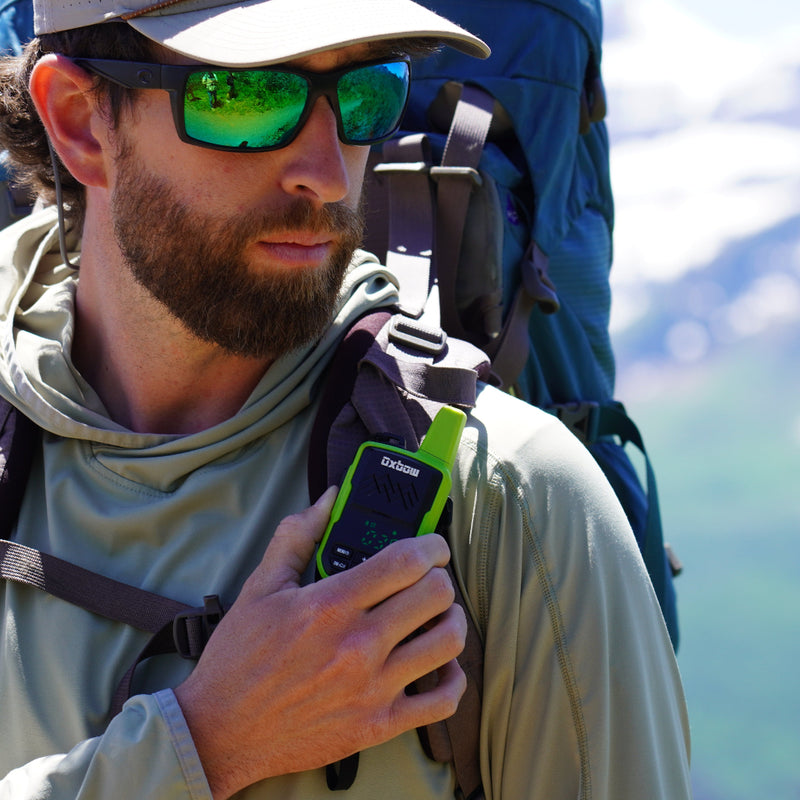 A bearded man wearing a beige cap, green mirrored sunglasses, and a light green hooded shirt packs a large blue hiking backpack while holding a green and black Garmin device outdoors against a blurred mountain background.