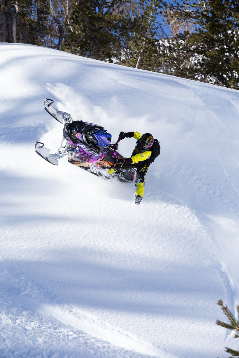 A snowmobile with a rider navigating through deep powder snow on a snowy slope, with evergreen trees and blue sky in the background.