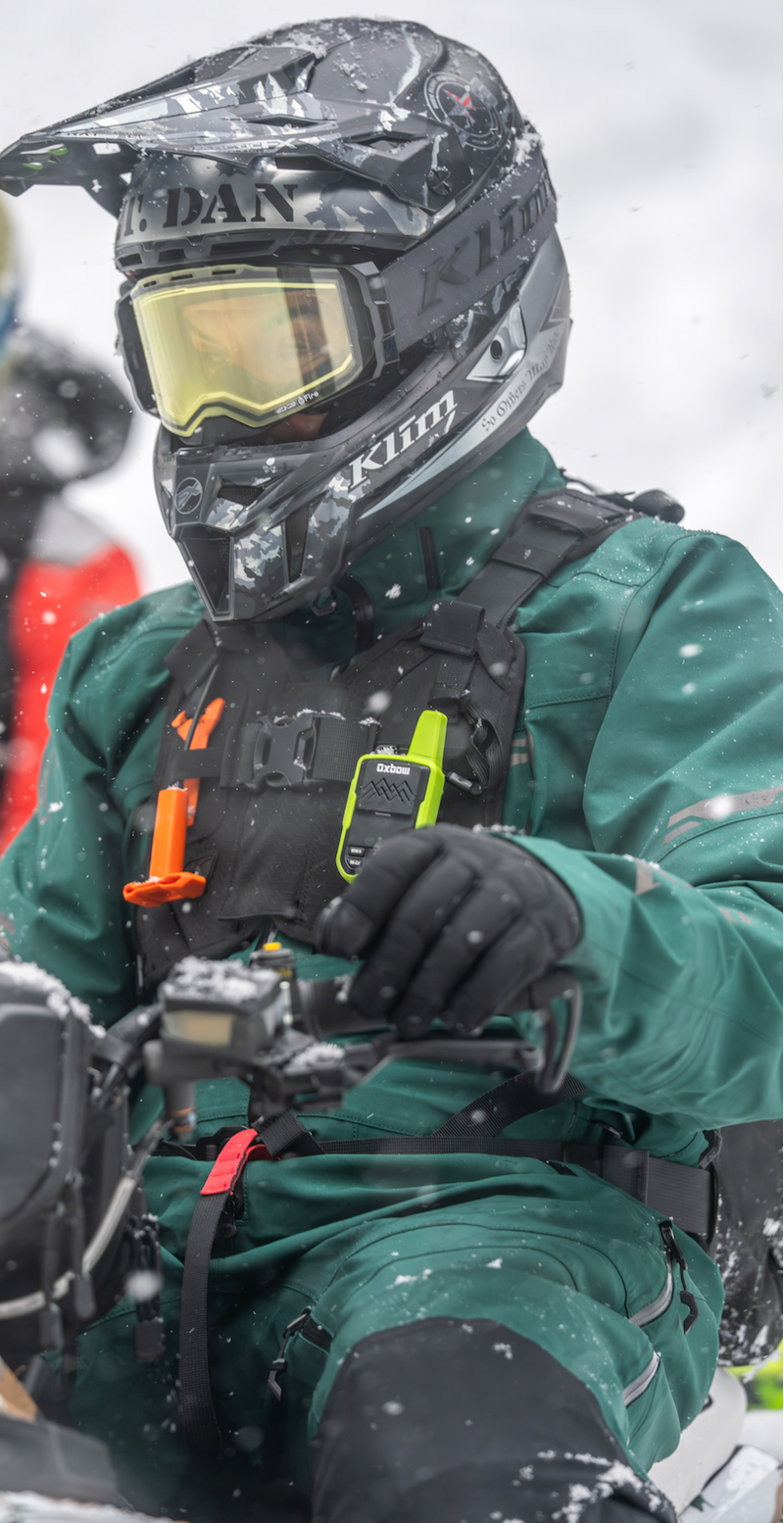 A person riding a snowmobile in the snow, wearing a black Klim helmet with a yellow lens, a green snowsuit, and a black harness with orange and lime green accents.